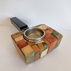 May include: A wooden espresso machine portafilter holder with a mosaic design. The holder is made of various wood tones and green resin, with a stainless steel portafilter basket. The handle is black.