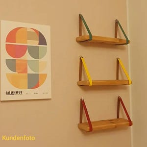 May include: Three rectangular wooden shelves mounted horizontally, each suspended by two colored leather straps in green, yellow, and red. Beside them hangs a rectangular poster featuring geometric semicircles in orange, blue, and tan with the text BAUHAUS.