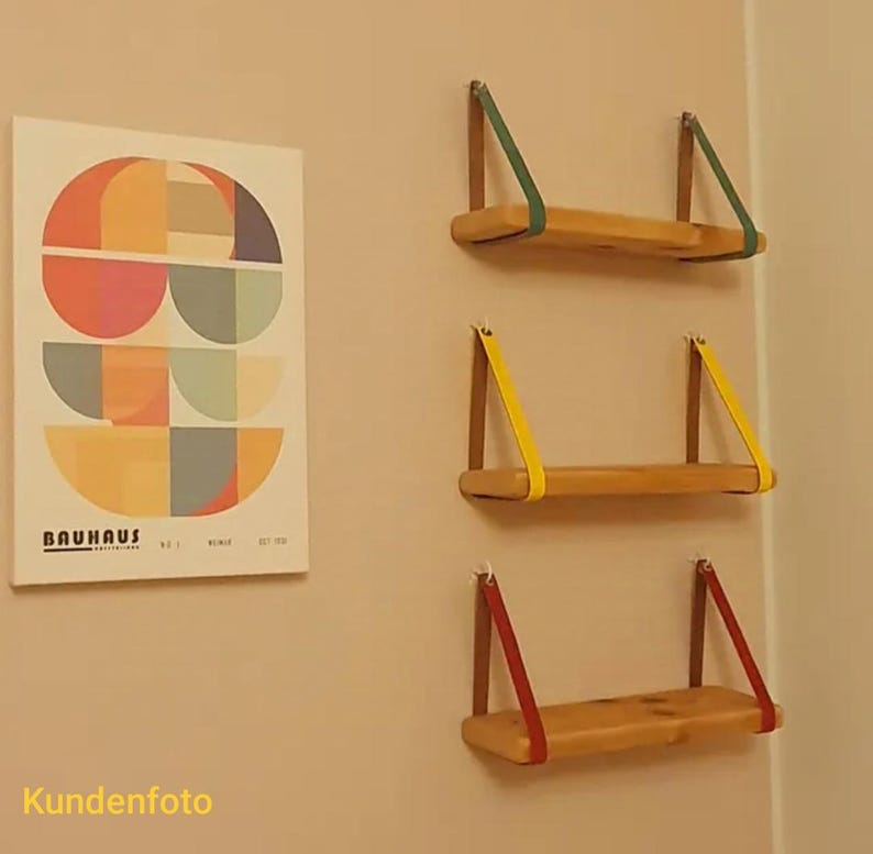 May include: Three rectangular wooden shelves mounted horizontally, each suspended by two colored leather straps in green, yellow, and red. Beside them hangs a rectangular poster featuring geometric semicircles in orange, blue, and tan with the text BAUHAUS.