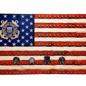 May include: A wooden wall display shaped like the United States flag with red, white, and blue painted stripes and a blue canton featuring the gold United States Coast Guard 1790 emblem. The stripes contain rows of small metal pegs for hanging items.