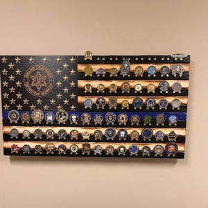 May include: Wooden American flag display with a blue line, holding numerous challenge coins. The flag features stars and stripes, with the words "Grand Traverse Detective Bureau" in a circular emblem. The coins are of various designs and colors.
