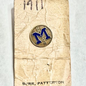 May include: Vintage gold-toned pin with a blue "M" emblem, set on a worn paper backing. The paper is marked with "1911" and the text "BURR, PATTERSON & AU.D CO., DETROIT". The pin is a collectible item.