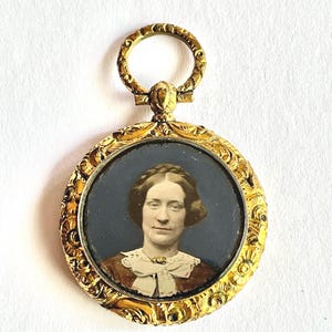 May include: A gold-toned antique pendant with a portrait of a woman. The round pendant features an ornate, filigree design around the portrait. The woman in the portrait is wearing a white collar and a brown dress. The pendant has a loop at the top.