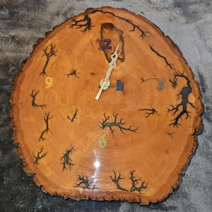 May include: A round wooden clock with a natural wood grain finish. The clock face features black and green tree-like designs and colorful numbers at the 3, 6, 9, and 12 positions. The clock hands are silver.