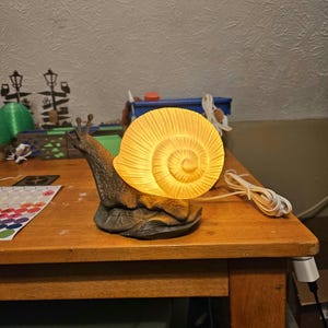 May include: A snail-shaped lamp featuring a dark gray, textured body and a translucent, glowing yellow shell with a spiral pattern. The lamp includes a white electrical cord and plug, emitting a warm light from the shell portion of the figure.