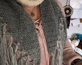 Könnte beinhalten: Ein grauer Häkel-Schal mit Fransen. Der Schal wird über einem rosafarbenen Pullover mit Schnürsenkel-Ausschnitt getragen.
