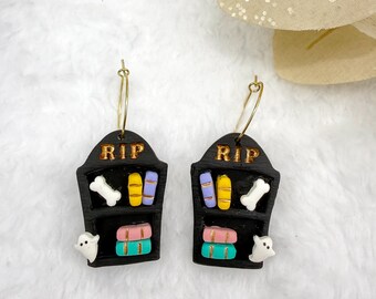 May include: A pair of Halloween-themed earrings. Each earring features a black tombstone design with the letters "RIP" in gold. The tombstones have shelves with colorful books, a bone, and a ghost.