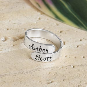 May include: A silver-colored bypass ring featuring two overlapping ends. The top end is stamped with the name Amber in a black script font, and the bottom end is stamped with the name Scott in a matching black script font. The metal band has a smooth, matte finish.