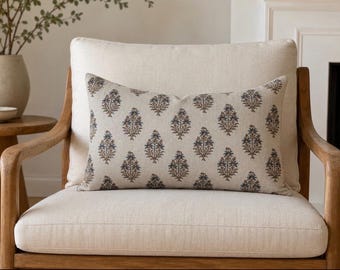May include: A rectangular throw pillow featuring a light beige fabric base with a repeating pattern of small, stylized floral motifs in shades of muted blue and brown. The pillow is positioned on a light-colored upholstered chair.