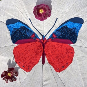 May include: A fabric butterfly quilt block with red, blue, and burgundy wings, set against a white background. The wings have detailed patterns. Two flowers are placed near the quilt. The text "Sewn by @off.kilter.quilter" is at the top.