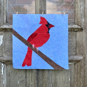 Może przedstawiać: Czerwony kardynał z czarną łatą na oku siedzący na brązowej gałęzi na jasnoniebieskim tle. Ptak jest wykonany z tkaniny i jest częścią bloku kołdry.