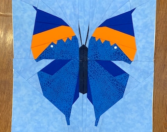 May include: A fabric butterfly quilt block with a blue, orange, and navy color scheme. The butterfly has detailed wings with a crackle pattern, and the background is a light blue. The block is on a wooden surface.