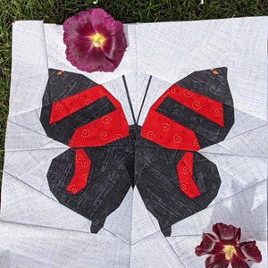 May include: A fabric butterfly quilt block with black and red wings, set against a white background. The wings have red stripes and a black body. The text "Sewn by @off.kilter.quilter" is at the top. Two dark purple flowers are placed near the quilt.