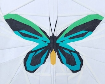 May include: A white square fabric block featuring a colorful butterfly design. The butterfly has teal, black, green, and yellow accents. The text "Sewn by @jbcmoom3" is at the bottom. This is a quilt block.