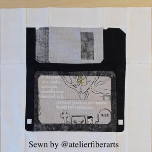 Op de afbeelding: Een stoffen blok met een zwart floppy disk-ontwerp met een grijs en wit interieur. Het interieur bevat tekst over een zombie-apocalyps. Het blok is wit omlijst met een ruitpatroon. De tekst onderaan luidt "Sewn by @atelierfiberarts."