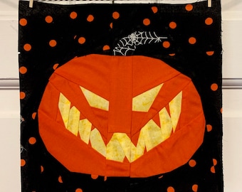 Op de afbeelding: Een zwarte stof met oranje stippen heeft een grote oranje jack-o'-lantern met een tandenrij en een wit spinnenweb erop.