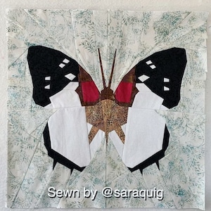 May include: A white butterfly with black wings and red accents is sewn from fabric. The butterfly is on a white background with a green floral pattern. The text "Sewn by @saraquig" is at the bottom of the image.
