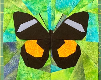 May include: A colorful butterfly quilt block with a black butterfly with orange and gray accents. The background is a green and blue fabric with a mottled pattern. The text "Sewn by @annalandesbenz" is at the bottom of the block.