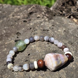May include: A bracelet made of gray, white, and brown agate beads with a large, light purple agate bead in the center. The bracelet is on a gray rock.