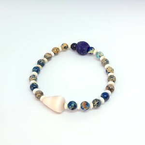 May include: A beaded bracelet featuring a variety of natural stone beads in shades of blue, tan, and white. The bracelet includes a large, light-colored shell-shaped bead and a large, dark purple bead. The bracelet is approximately 7 inches in circumference.