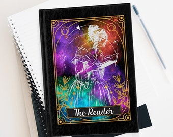 Puede incluir: Una libreta negra con un marco dorado y un fondo de galaxia colorido. La libreta presenta una silueta blanca de una persona leyendo un libro con el texto "The Reader" en la portada.
