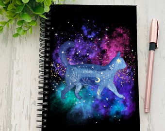 Puede incluir: Una libreta negra de espiral con un diseño de galaxia que presenta una silueta de gato azul con estrellas doradas y símbolos celestiales. El gato está rodeado por una nebulosa de color rosa, morado y verde.