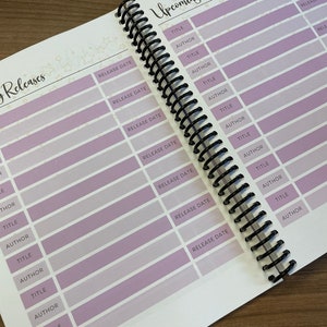 Puede incluir: Primer plano de un libro con un dise&ntilde;o morado y blanco. El libro tiene una encuadernaci&oacute;n en espiral y est&aacute; abierto en una p&aacute;gina con una lista de lanzamientos pr&oacute;ximos. La lista incluye columnas para el t&iacute;tulo, el autor y la fecha de lanzamiento.