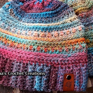 Pu&ograve; includere: Due cappelli all'uncinetto colorati con un'etichetta in pelle marrone che dice "Nana's Crochet Creations" e un'icona di filato.
