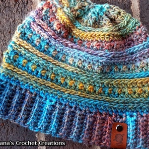 May include: A colourful crocheted beanie hat with a brown leather tag that says "Nana's Crochet Creations". The hat has a blue, green, yellow, and purple colour scheme.