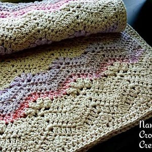 May include: A crocheted blanket with a beige background and stripes of pink, purple, and lavender. The blanket is folded in half, with the top half of the blanket folded over the bottom half. The blanket is made with a simple stitch pattern and has a soft, textured feel.