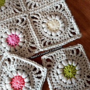 May include: Close-up of a crocheted blanket with a repeating pattern of flowers in pink, white, and green. The blanket is made of a light gray yarn and has a textured surface.