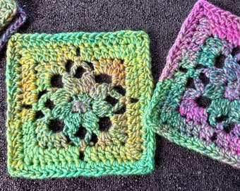 May include: Two crocheted squares with a floral design in shades of green, yellow, pink, and blue. The squares are made with a variegated yarn and have a textured, lacy look.