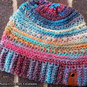 Pu&ograve; includere: Un berretto lavorato a crochet colorato con un design sfumato multicolore. Il berretto ha un motivo a punto testurizzato e un piccolo cartellino in pelle marrone con un design a uncinetto e filo. Il cartellino riporta il testo "Nana's Crochet Creations".