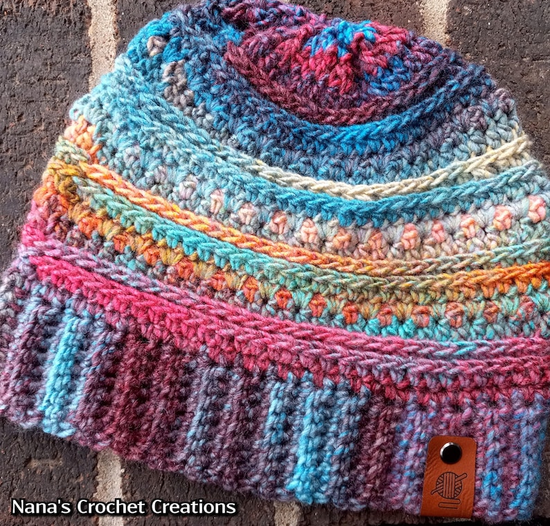 Pu&ograve; includere: Un berretto lavorato a crochet colorato con un design sfumato multicolore. Il berretto ha un motivo a punto testurizzato e un piccolo cartellino in pelle marrone con un design a uncinetto e filo. Il cartellino riporta il testo "Nana's Crochet Creations".