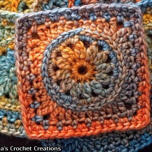 May include: A close-up of a crocheted square with a circular design in the center. The square is made with a variety of shades of blue, orange, and brown yarn.