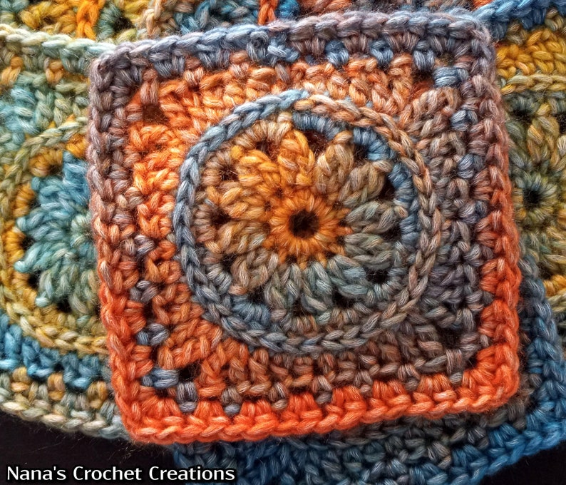 May include: A close-up of a crocheted square with a circular design in the center. The square is made with a variety of shades of blue, orange, and brown yarn.