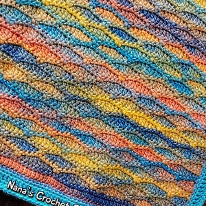 May include: A crocheted blanket featuring a diagonal wave pattern in variegated shades of yellow, orange, blue, and green. The fabric has a textured surface with a solid turquoise border. The text Nana's Crochet Creations is printed in white.