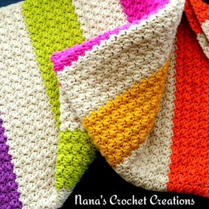 Könnte beinhalten: Eine bunte gestreifte Häkeldecke in verschiedenen Farben wie Lila, Grün, Gelb, Orange und Pink. Die Decke ist mit einem strukturierten Stich gearbeitet und zur Hälfte gefaltet.