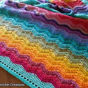 Może przedstawiać: Kolorowy koc szydełkowy z wzorem chevron w odcieniach fioletu, niebieskiego, zielonego, żółtego, pomarańczowego i czerwonego. Koc ma turkusową obwódkę z szydełkowym wykończeniem.