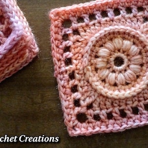 May include: A pink and white crochet square with a detailed flower design in the centre. The square is made with a gradient yarn that transitions from pink to white.