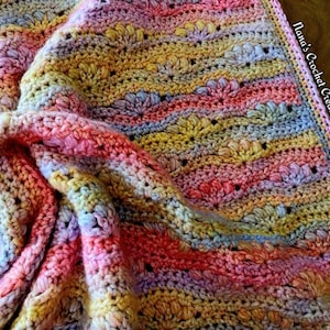Peut inclure: Une couverture au crochet faite à la main, colorée, avec un motif ondulé. La couverture présente un mélange de couleurs pastel, notamment du rose, du jaune, du bleu et du violet. La couverture est douce et texturée, avec le texte "Nana's Crochet Creations" visible sur le bord.