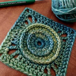 May include: A green and blue crochet square with a circular design in the center. The square has a textured, lacy pattern around the edges.