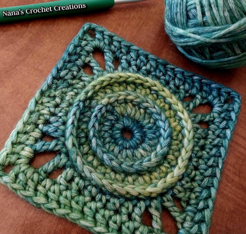 May include: A green and blue crochet square with a circular design in the center. The square has a textured, lacy pattern around the edges.