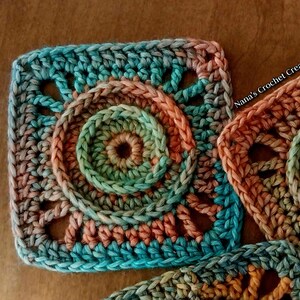 May include: A close-up of a crocheted square with a circular pattern in shades of teal, green, and peach. The square is part of a larger crocheted project.