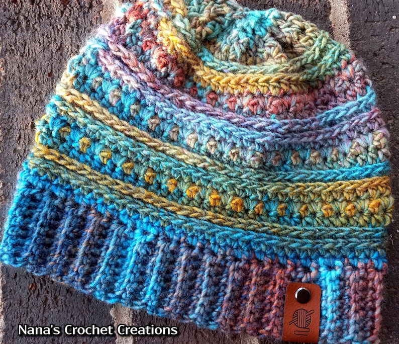 Pu&ograve; includere: Un berretto lavorato a uncinetto colorato con un'etichetta in pelle marrone che dice "Nana's Crochet Creations". Il berretto ha una combinazione di colori blu, verde, giallo e viola.