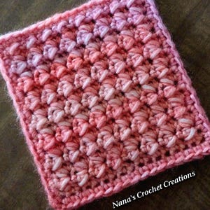 May include: A pink and white crochet square with a gradient pattern. The square is made with a textured stitch and has a border around the edges. The text "Nana's Crochet Creations" is visible in the bottom right corner of the image.