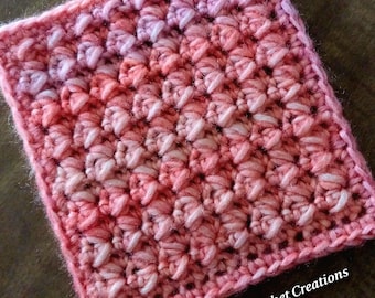 May include: A pink and white crochet square with a gradient pattern. The square is made with a textured stitch and has a border around the edges. The text "Nana's Crochet Creations" is visible in the bottom right corner of the image.