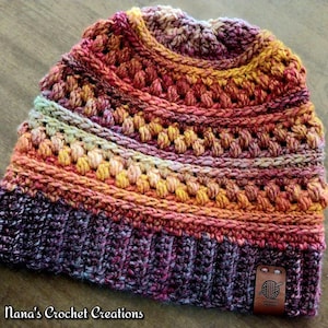 Può includere: Un berretto all'uncinetto fatto a mano in una miscela di colori tra cui viola, arancione, giallo e rosa. Il cappello presenta un bordo a coste e un motivo a punto strutturato. Una piccola etichetta in pelle marrone con un logo di filo e gancio è attaccata al bordo. Il testo "Nana's Crochet Creations" è in basso.