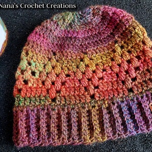May include: A colorful crocheted beanie hat with a brown leather tag that says "Nana's Crochet Creations". The hat has a multi-colored yarn with shades of pink, orange, green, and purple.