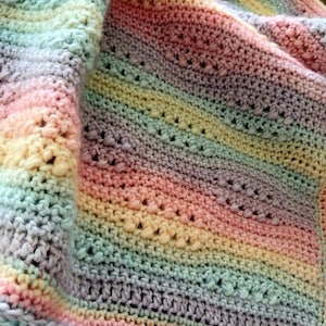 May include: A handmade crochet blanket featuring a soft pastel rainbow gradient of pink, yellow, blue, and purple yarn. The textile displays a textured pattern with alternating rows of solid stitches and openwork details. Text reads Nana's Crochet Creations.